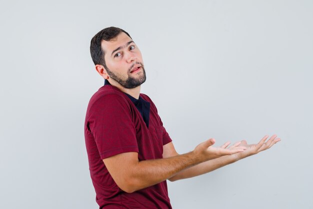 Young man in red t-shirt showing helpless gesture and looking troubled , front view.