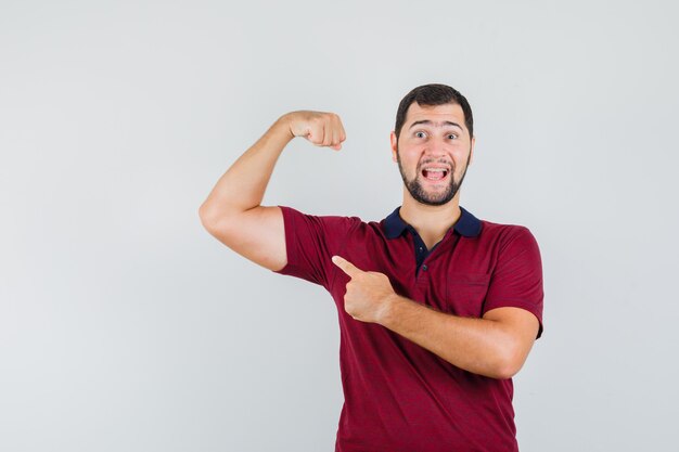 Молодой человек в красной футболке показывает мышцы рук и выглядит веселым, вид спереди.