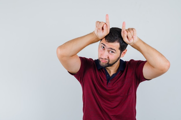 Young man in red t-shirt showing horn sign over his head and looking weird , front view.