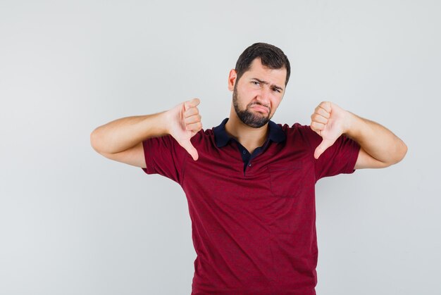 Young man in red t-shirt showing thumb down and looking displeased , front view.