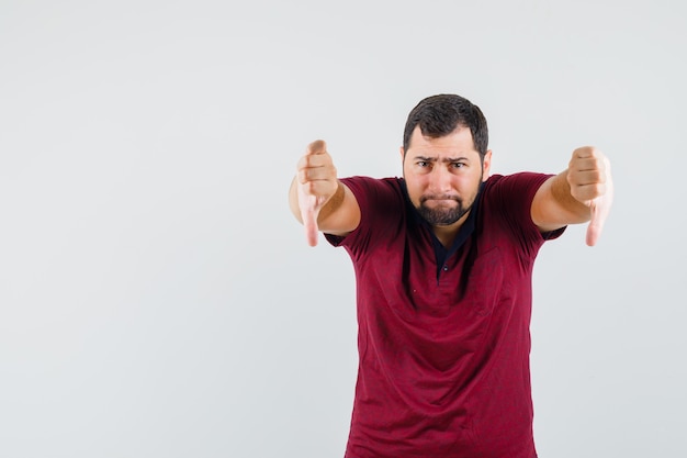 Free photo young man in red t-shirt showing thumb down and looking embarrassed , front view.