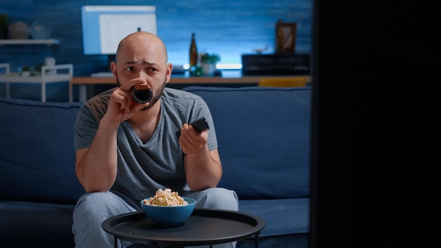 Young man relaxing on cozy couch, eating popcorn and drinking beer using remote controller switching tv channels, searching funny online film movie, spending free weekend time alone at home.
