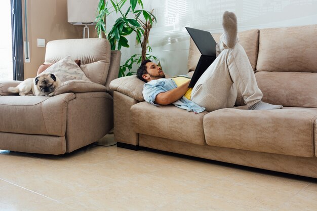 Young man resting on a sofa at home and using a laptop with his dog beside him