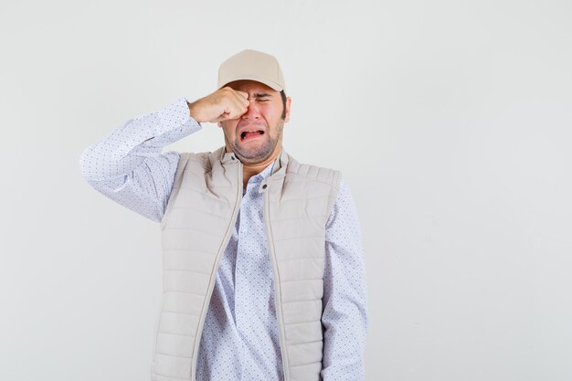 Young man rubbing eye and crying in beige jacket and cap and looking lugubrious. front view.