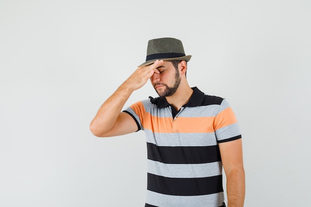 Young man rubbing eyes and nose in t-shirt, hat and looking tired