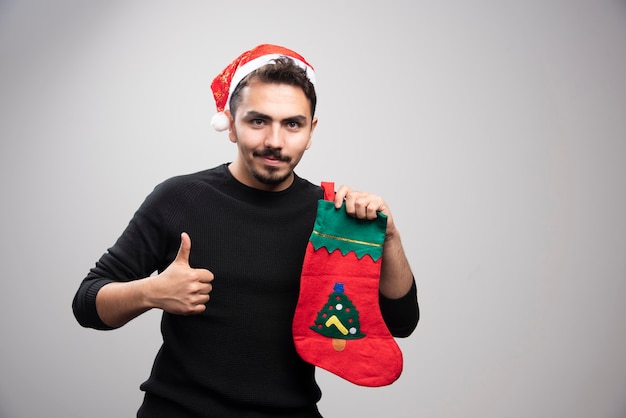 A young man in a Santa's hat showing a thumb up and holding a Christmas sock .