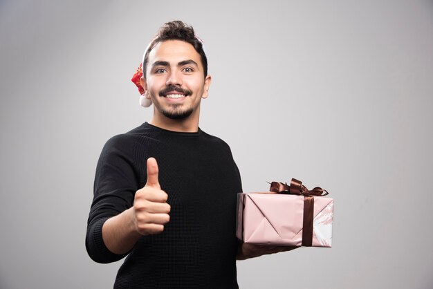 A young man in a Santa's hat showing a thumb up and holding a gift .