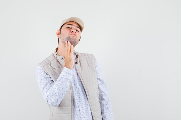 Young man scratching chin in beige jacket and cap and looking focused , front view.