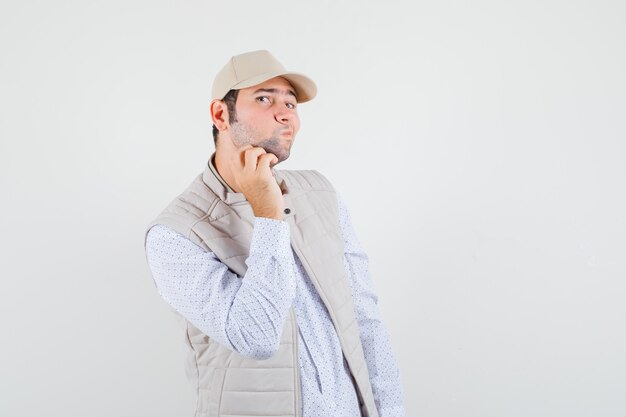 Young man scratching face in beige jacket and cap and looking focused. front view.