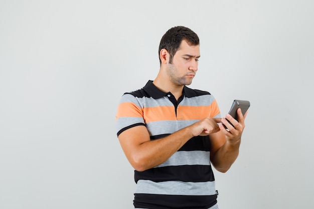 Young man searching something at phone in t-shirt and looking concentrated. front view.