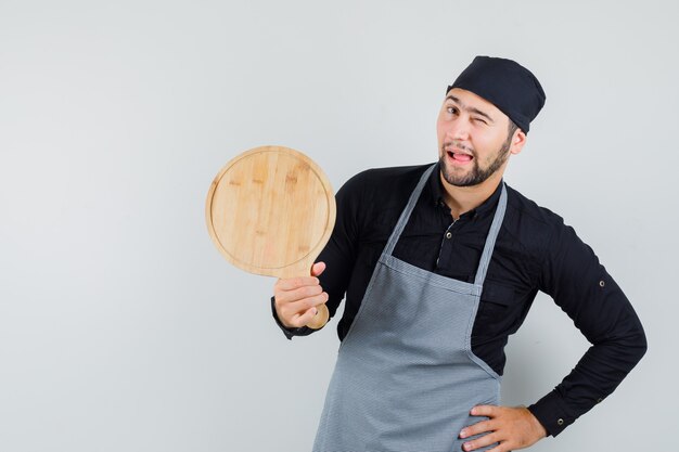 Young man in shirt, apron holding cutting board and winking eye , front view.