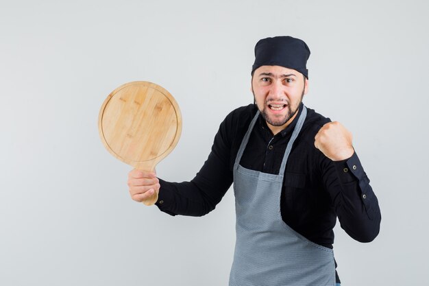 Young man in shirt, apron holding cutting board with raised fist and looking confident , front view.