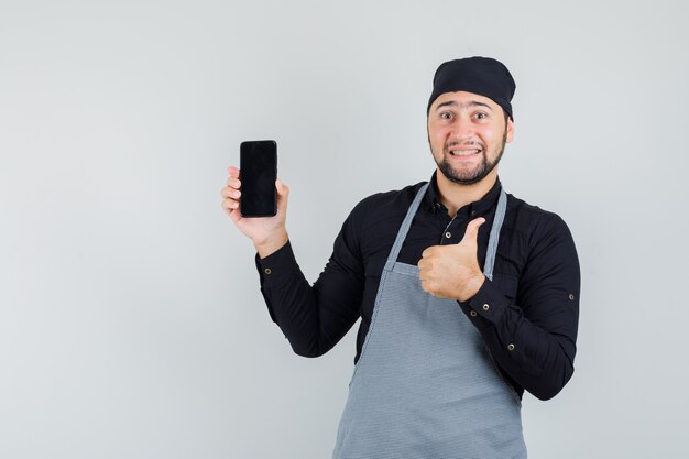 Young man in shirt, apron holding mobile phone with thumb up and looking glad , front view.