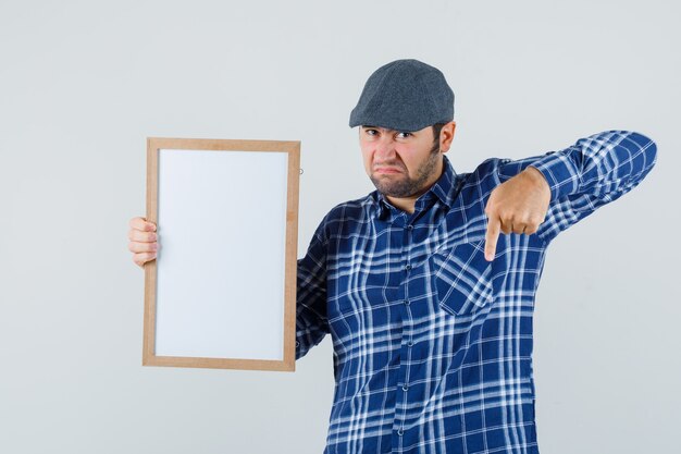 Young man in shirt, cap holding empty frame, pointing down and looking doubtful , front view.