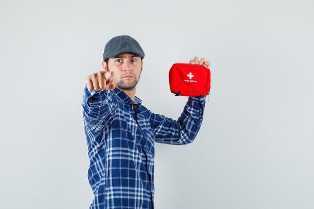 Young man in shirt, cap pointing at camera, holding first aid kit and looking serious .