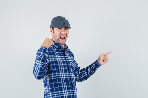 Young man in shirt, cap pointing to the side and looking blissful , front view.