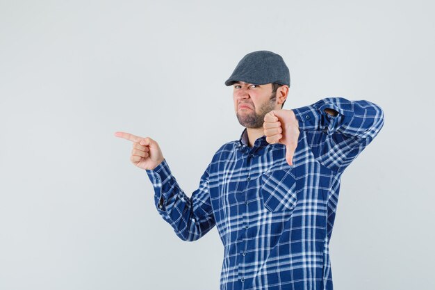 Young man in shirt, cap pointing to the side, showing thumb down and looking discontent , front view.