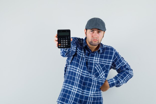 Young man in shirt, cap showing calculator and looking doubtful , front view.