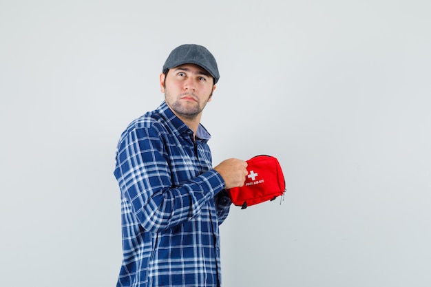 Young man in shirt, cap trying to open first aid kit and looking hesitant , front view.