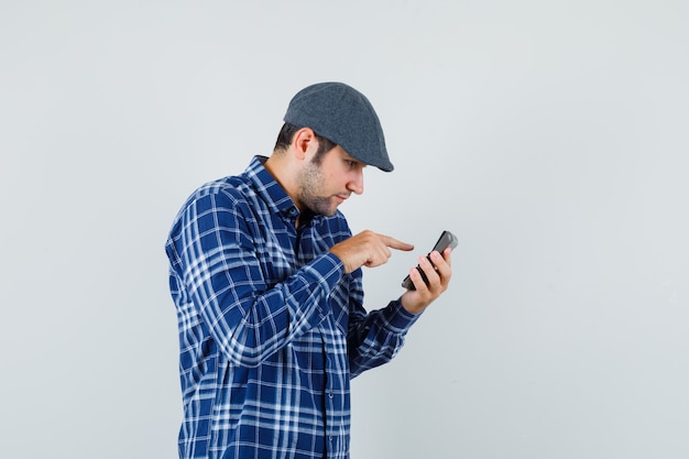 Giovane uomo in camicia, berretto utilizzando la calcolatrice e guardando occupato, vista frontale.