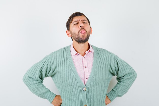 Young man in shirt, cardigan keeping hands on waist, sticking tongue out and looking grumpy , front view.