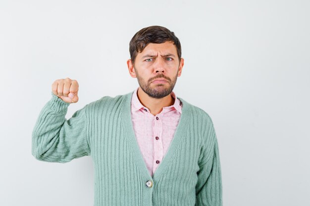 Young man in shirt, cardigan threatening with fist and looking serious , front view.