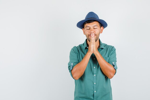 Young man in shirt, hat holding hands in praying gesture and looking hopeful