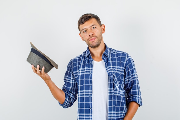 Young man in shirt holding hat and looking careful, front view.