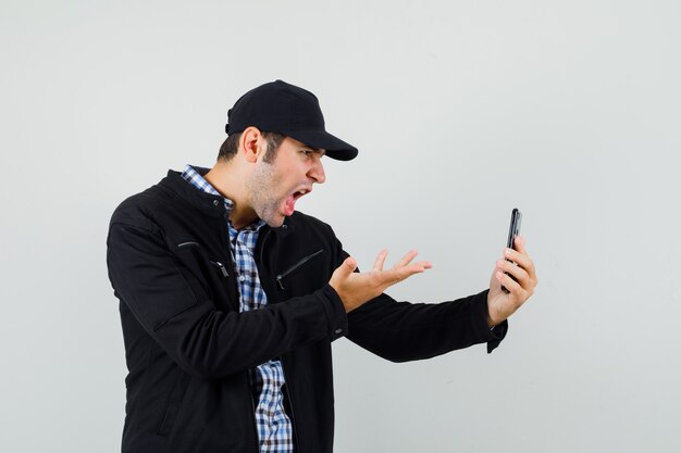 Young man in shirt, jacket, cap discussing something on video chat and looking angry