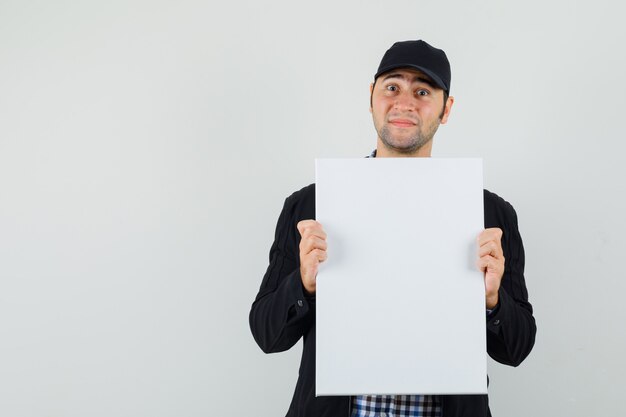 Young man in shirt, jacket, cap holding blank canvas and looking optimistic