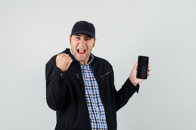 Young man in shirt, jacket, cap holding mobile phone, showing winner gesture and looking happy