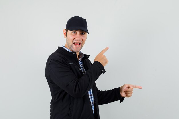 Young man in shirt, jacket, cap pointing fingers away and looking cheery