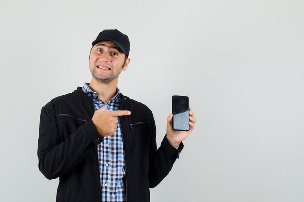 Young man in shirt, jacket, cap pointing at mobile phone and looking merry