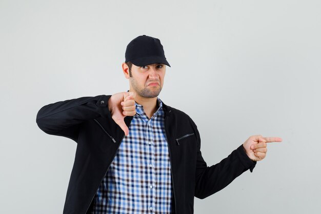 Young man in shirt, jacket, cap pointing to the side, showing thumb down and looking displeased