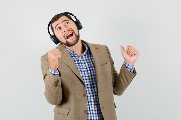 Young man in shirt, jacket enjoying music with headphones and looking merry , front view.