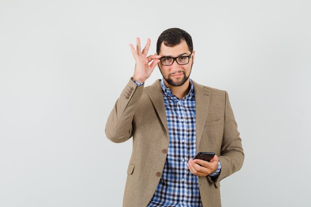 Young man in shirt, jacket holding mobile phone, touching glasses and looking confident , front view.