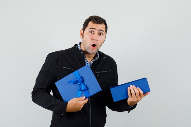 Молодой человек в рубашке, куртке держит открытую подарочную коробку и выглядит изумленным