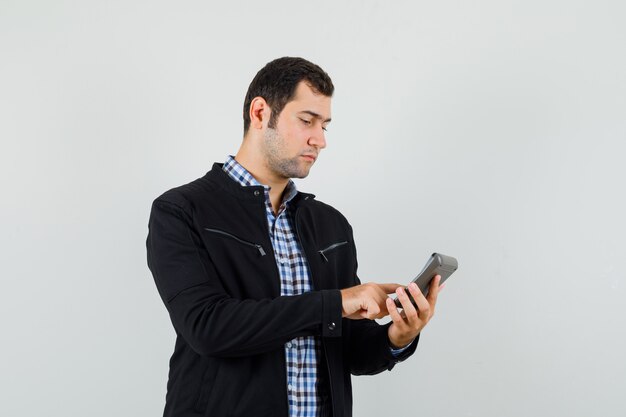 Young man in shirt, jacket making calculations on calculator and looking busy