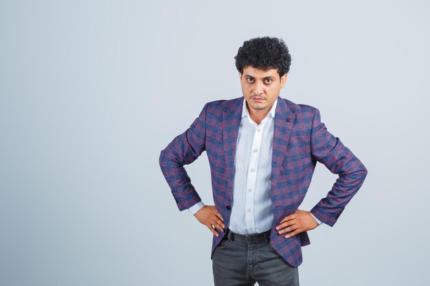 Young man in shirt, jacket, pants keeping hands on waist and looking angry , front view.