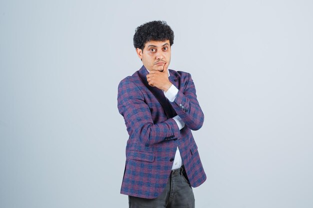 Young man in shirt, jacket, pants propping chin on hand and looking pensive , front view.