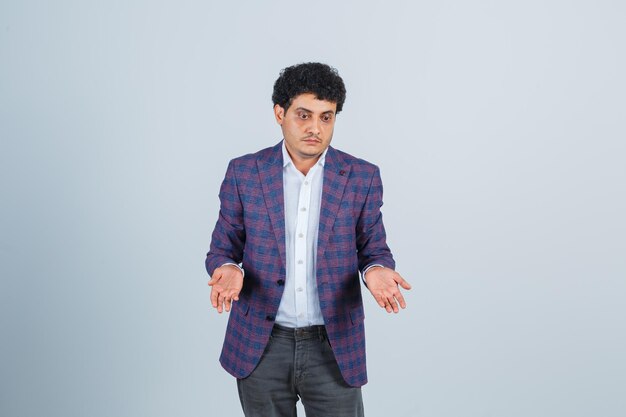 Young man in shirt, jacket, pants showing helpless gesture and looking confused , front view.