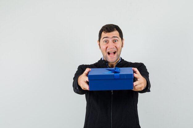 Young man in shirt, jacket presenting gift box and looking merry