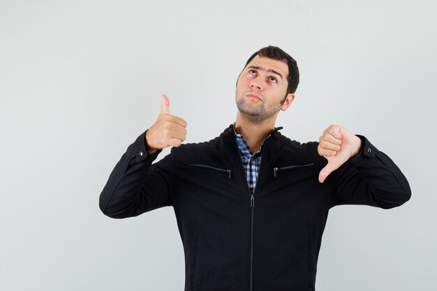 Young man in shirt, jacket showing thumbs up and down and looking irresolute