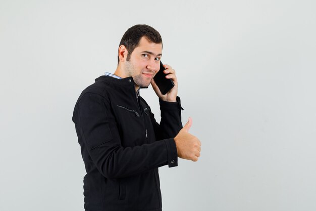 Молодой человек в рубашке, куртке разговаривает по мобильному телефону, показывает большой палец вверх и выглядит весело