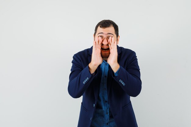 Young man in shirt, jacket telling secret with hands near mouth , front view.