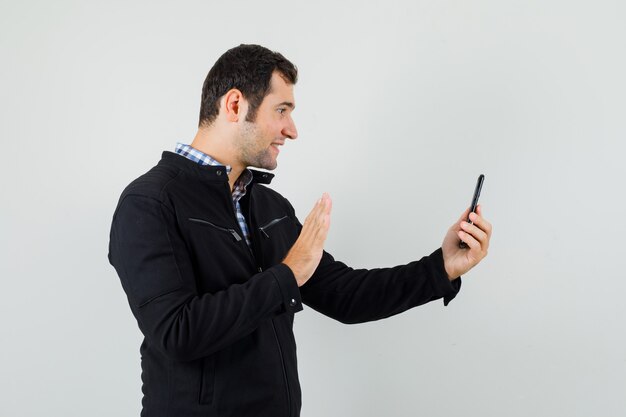 Young man in shirt, jacket waving hand on video chat and looking cheery