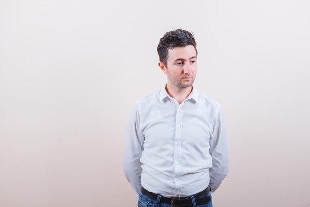 Young man in shirt, jeans looking away, holding hands behind back and looking concerned