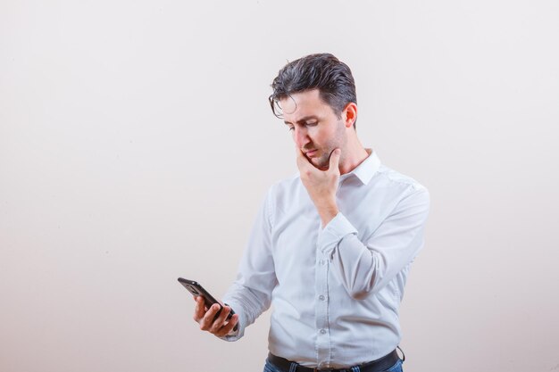 Young man in shirt, jeans looking at mobile phone and looking pensive