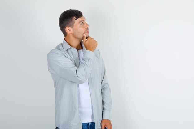 Young man in shirt, jeans looking up with hand on chin