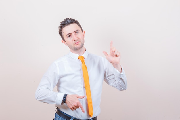 Young man in shirt, jeans pointing fingers up and down and looking irresolute